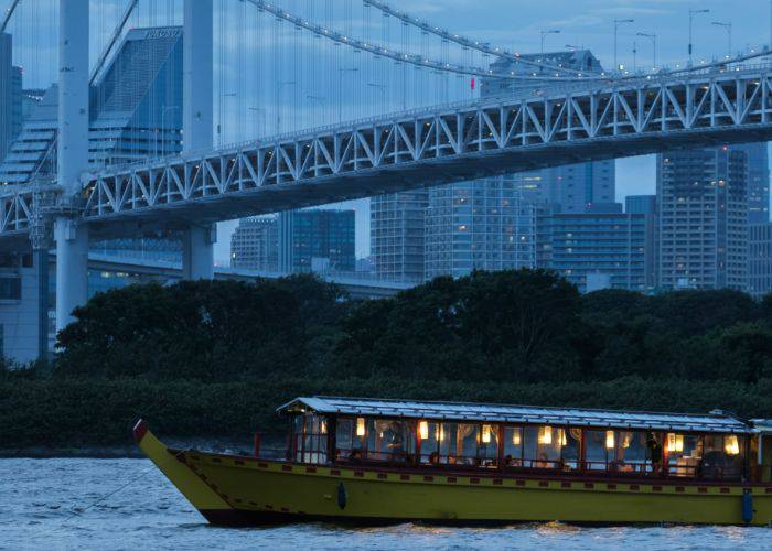 The Funasei Yakatabune, a yellow and red traditional Japanese pleasure boat traveling under Rainbow Bridge.
